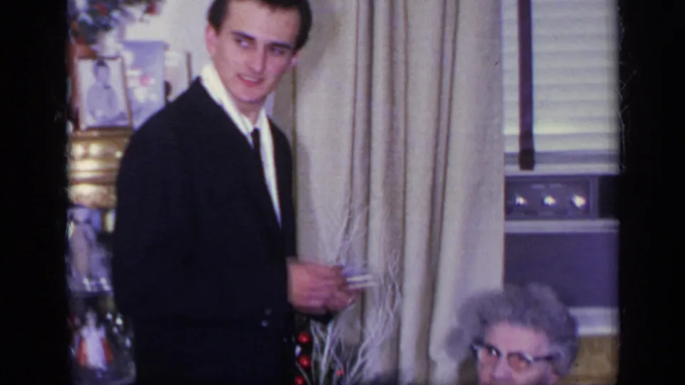 1969 California home-movie frame of a suited young man near a window, with an older woman partly visible at the lower edge.