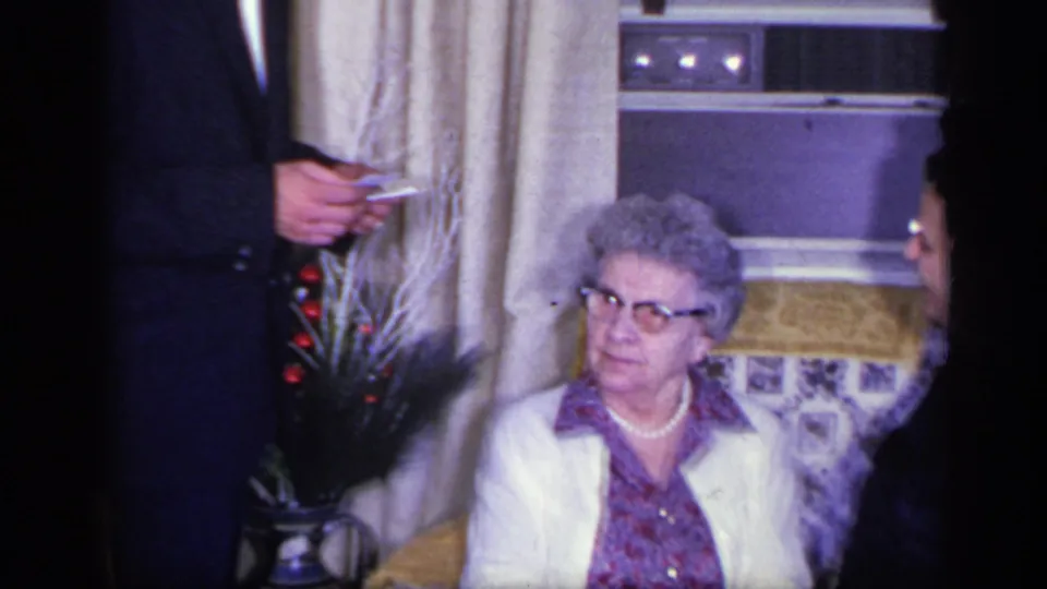 1969 California home-movie frame of an older woman seated on a sofa as a suited man and another person are partly visible nearby.