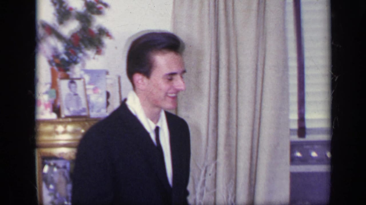 Young man in a dark suit smiles indoors beside curtains and framed tabletop photos.