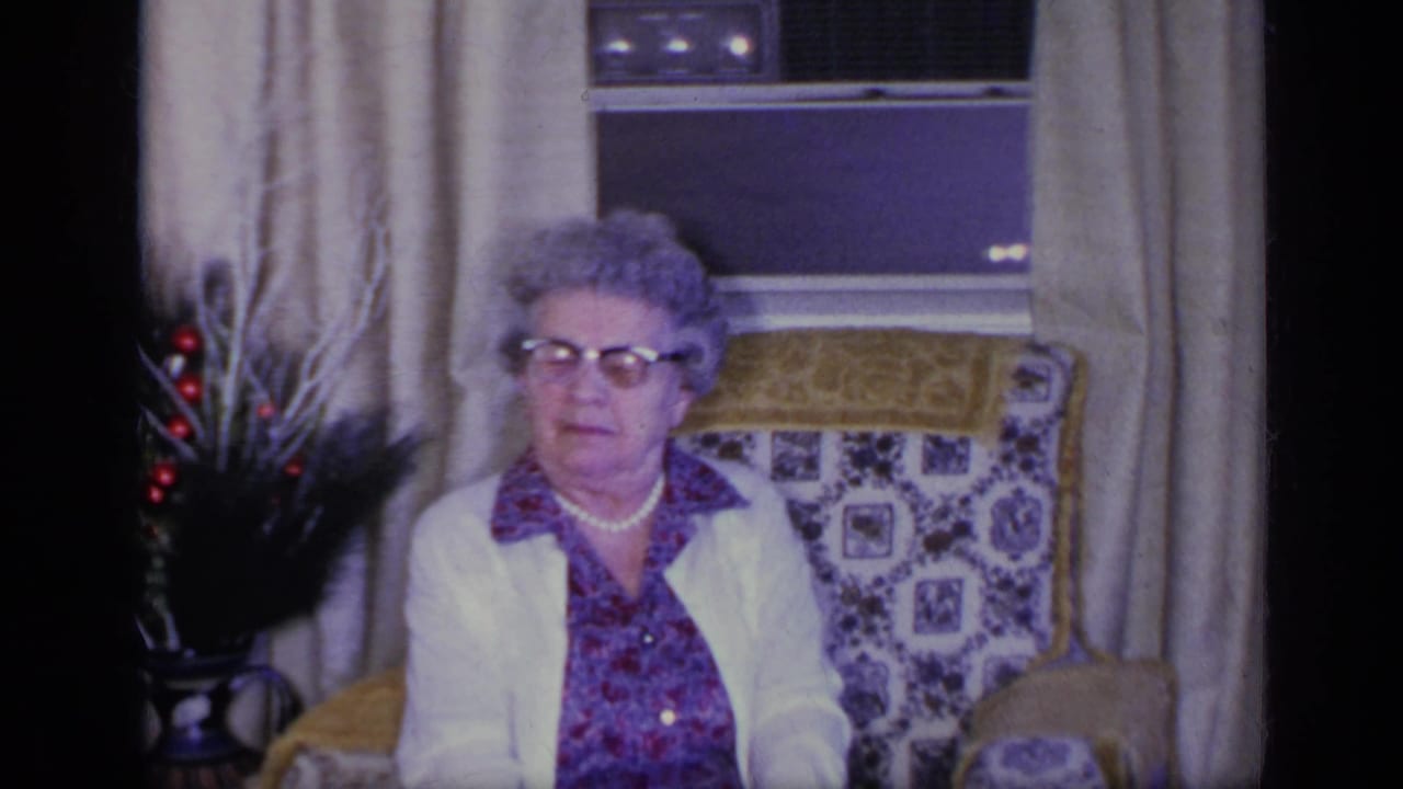 Elderly woman with glasses sits in a patterned armchair near curtains and a vase.