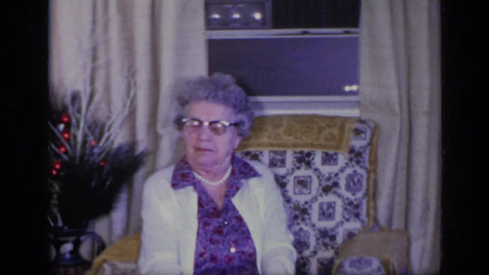 An elderly woman wearing glasses and a white cardigan sits in a patterned chair in a California living room, 1969.
