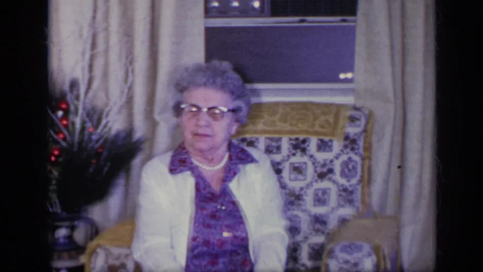 The older woman remains seated in the living room, framed by curtains, a window, and patterned upholstery in 1969 home-movie footage.