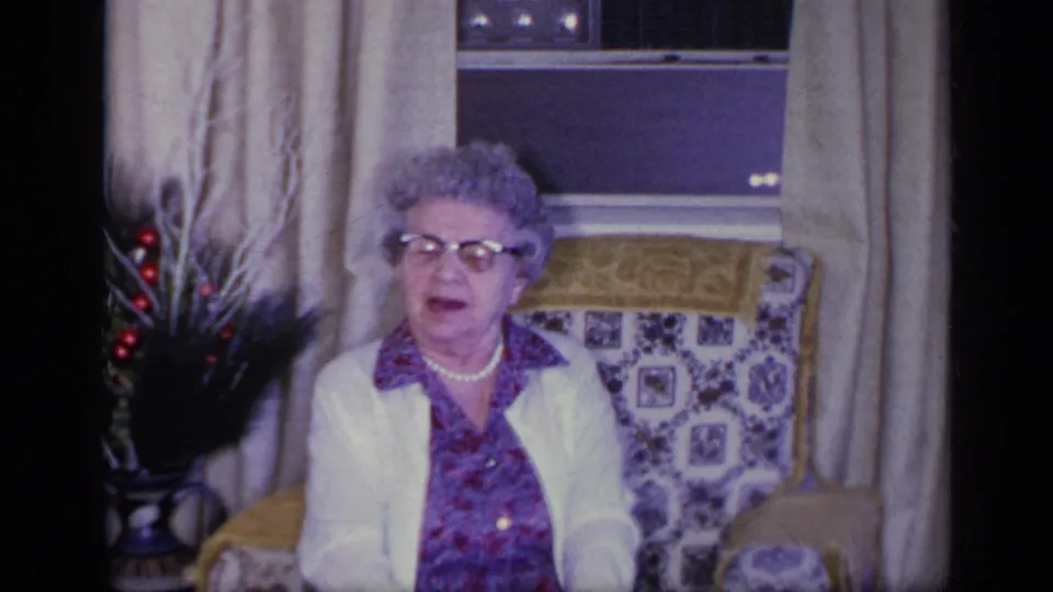 In the 1969 California living room scene, the elderly woman appears to speak while seated near curtains, a window, and a decorative vase.