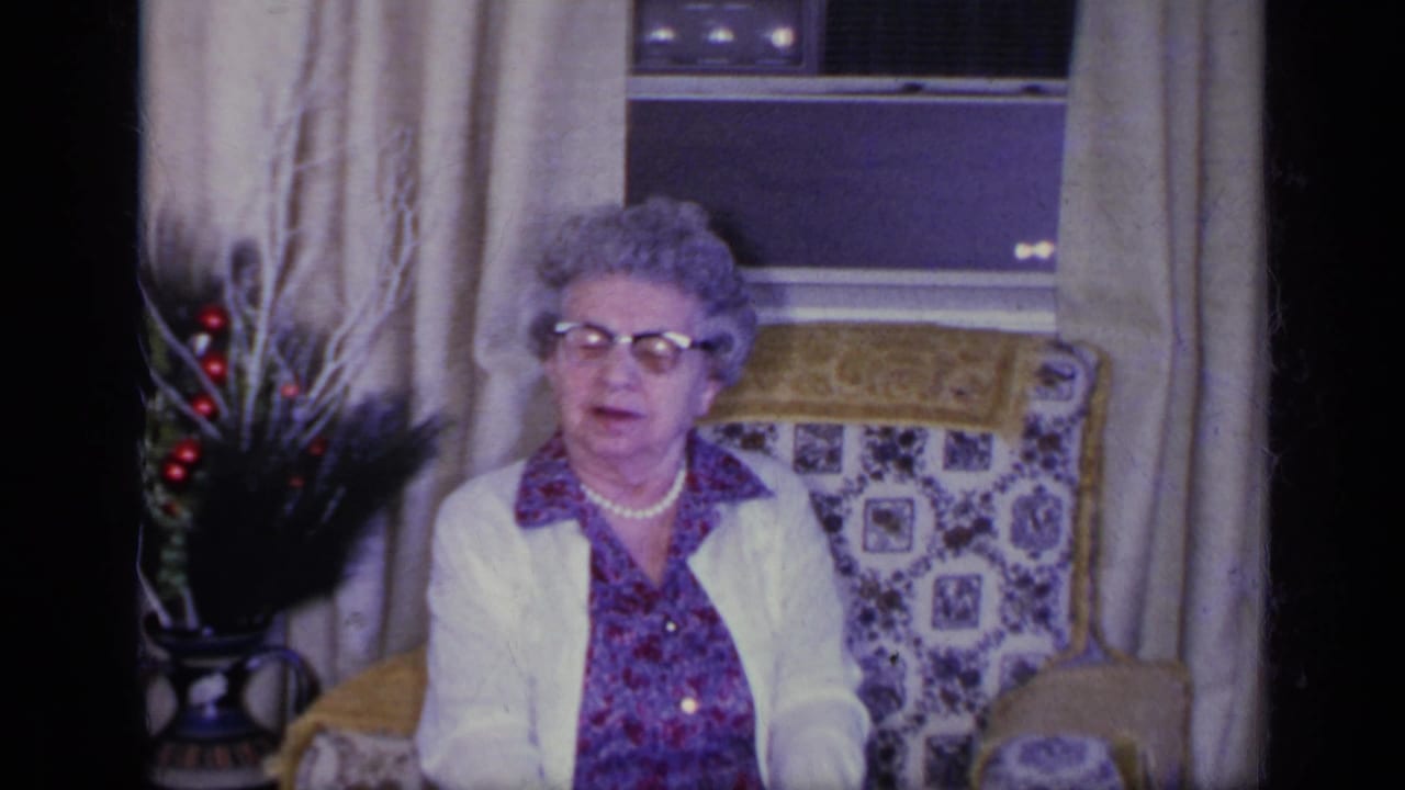 Older woman in glasses seated in a patterned armchair beside drapes and a window.