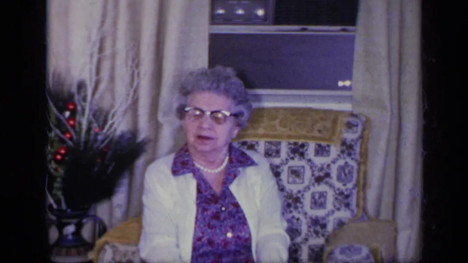 1969 California home-movie frame showing an older woman in glasses, blouse and white cardigan seated by a draped window.