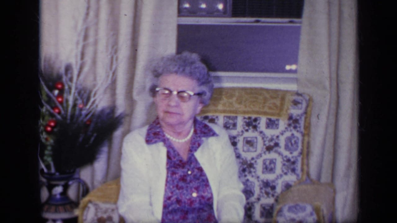 Older woman seated by a window with a decorative plant visible beside the chair.