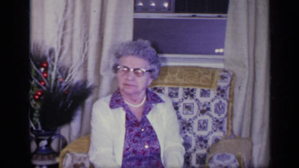 1969 California home-movie frame of an older woman in an armchair, with beige drapes, window and decorative plant nearby.
