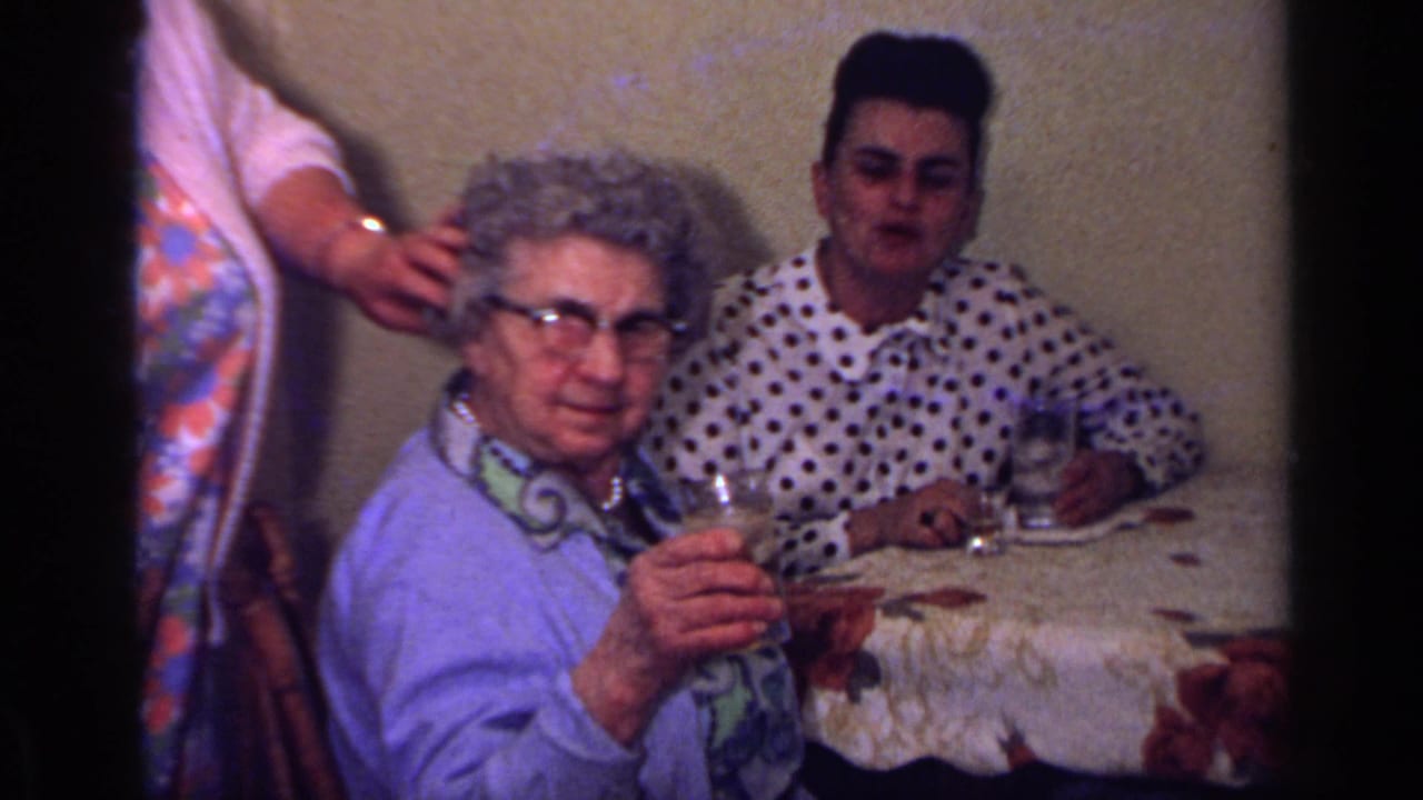 1969 California home-movie frame of an older woman in a light blue cardigan raising a glass at an indoor family table.