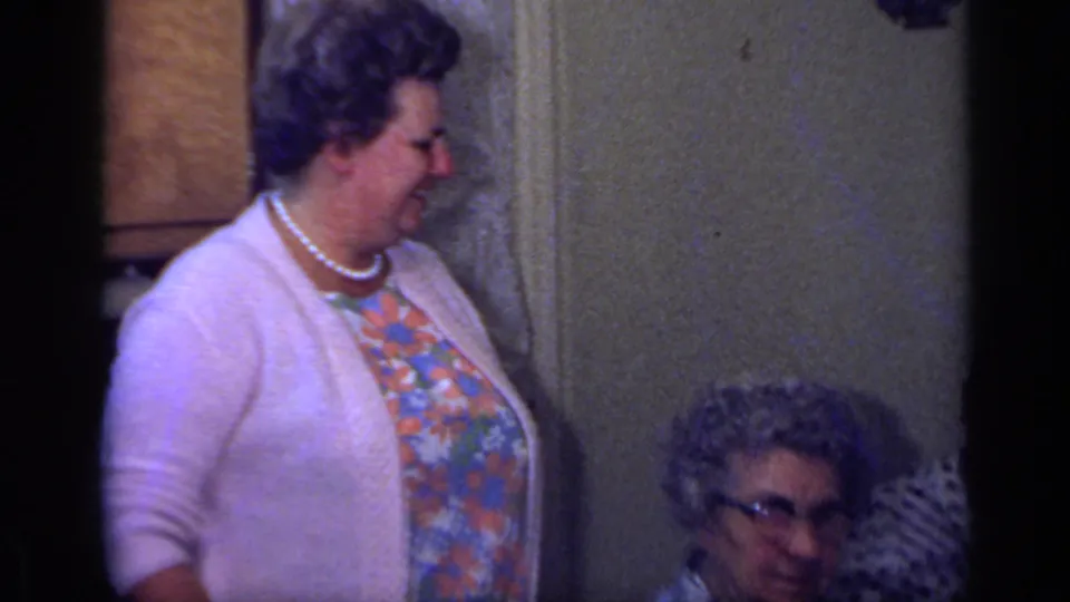 1969 California home-movie frame showing a woman in pearls standing near the older woman at a family celebration.