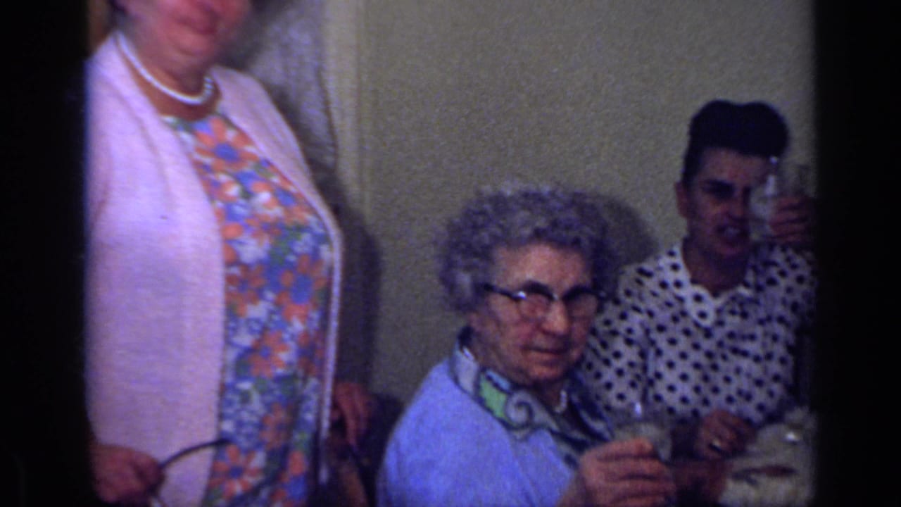 Elderly woman with glasses sits at a table while a young man sits behind her and a woman stands nearby.