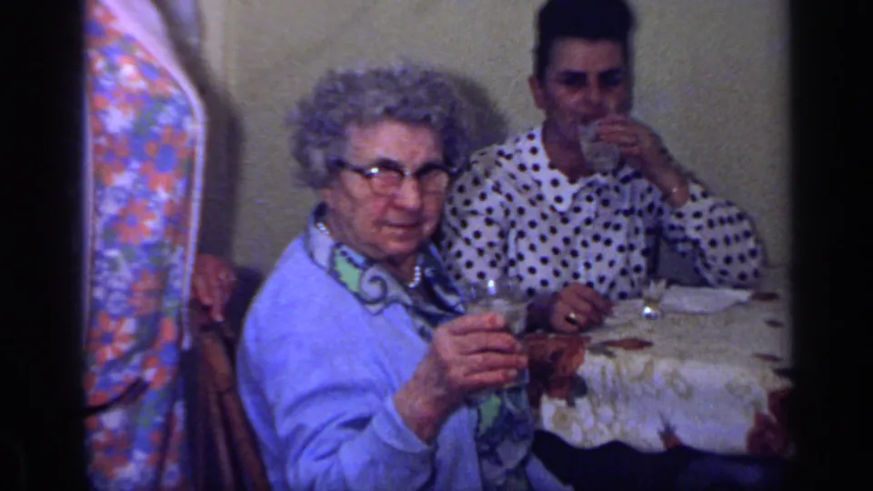 1969 California home-movie frame of an elderly woman raising a drink at a table, with a young man in a polka-dot shirt seated behind her.