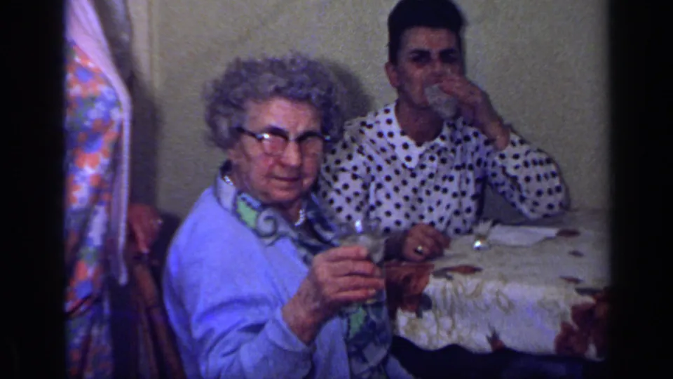 1969 California 8mm frame showing an elderly woman at a table holding a glass as a young man behind her drinks from his own glass.