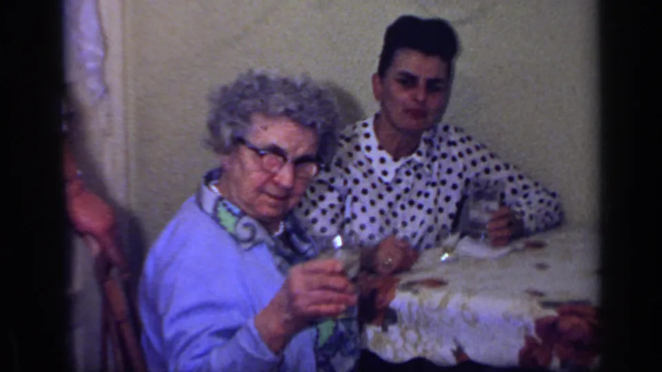 1969 California home-movie frame of an elderly woman in a blue cardigan and a young man in a polka-dot shirt seated with drinks at a table.