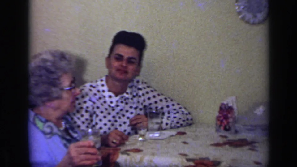 In a 1969 California home-movie frame, two people sit at a dining table with drinks, a floral tablecloth, and a plain wall behind them.