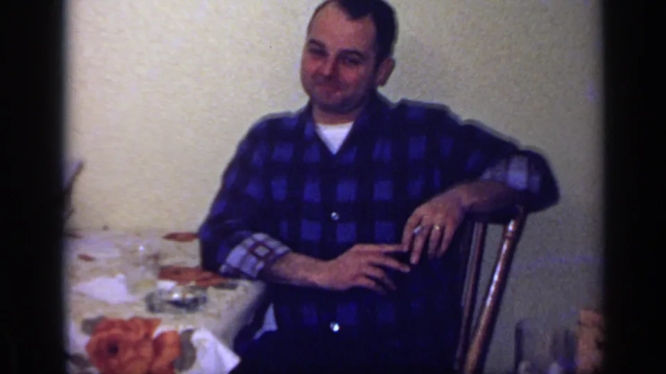A man in a blue checked shirt sits at a dining table in a 1969 California home interior, smiling with a cigarette in hand.