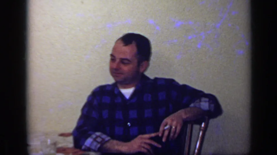 The same man sits back beside the dining table in a 1969 California home-movie frame, with a glass and table edge visible nearby.