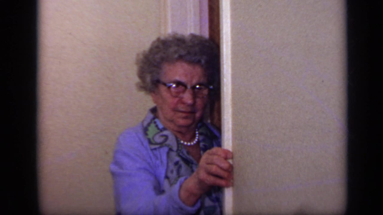 Older woman with glasses peeking from behind an interior doorframe.