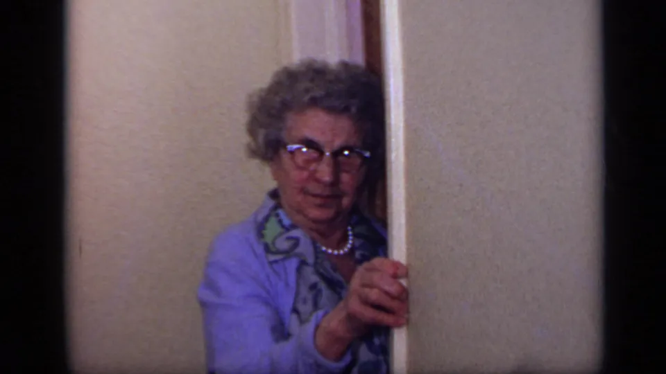 The older woman remains partly hidden behind the interior doorframe, holding it as she looks outward.