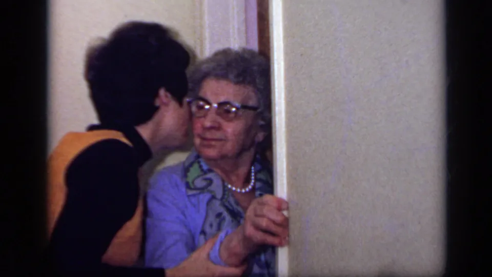 A younger woman in an orange vest leans toward the older woman at the doorway for a cheek kiss.