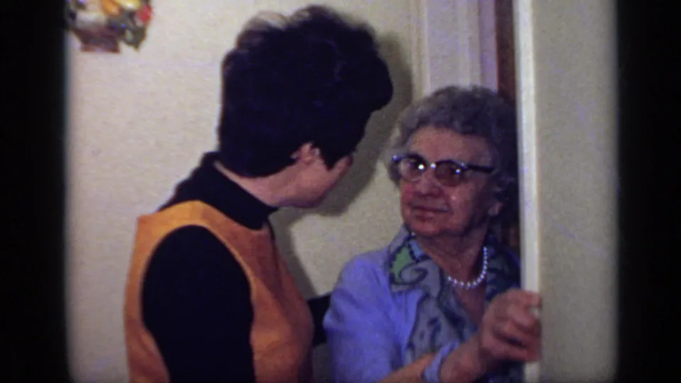The younger woman stands close to the older woman after the greeting, both framed by the doorway and wall.