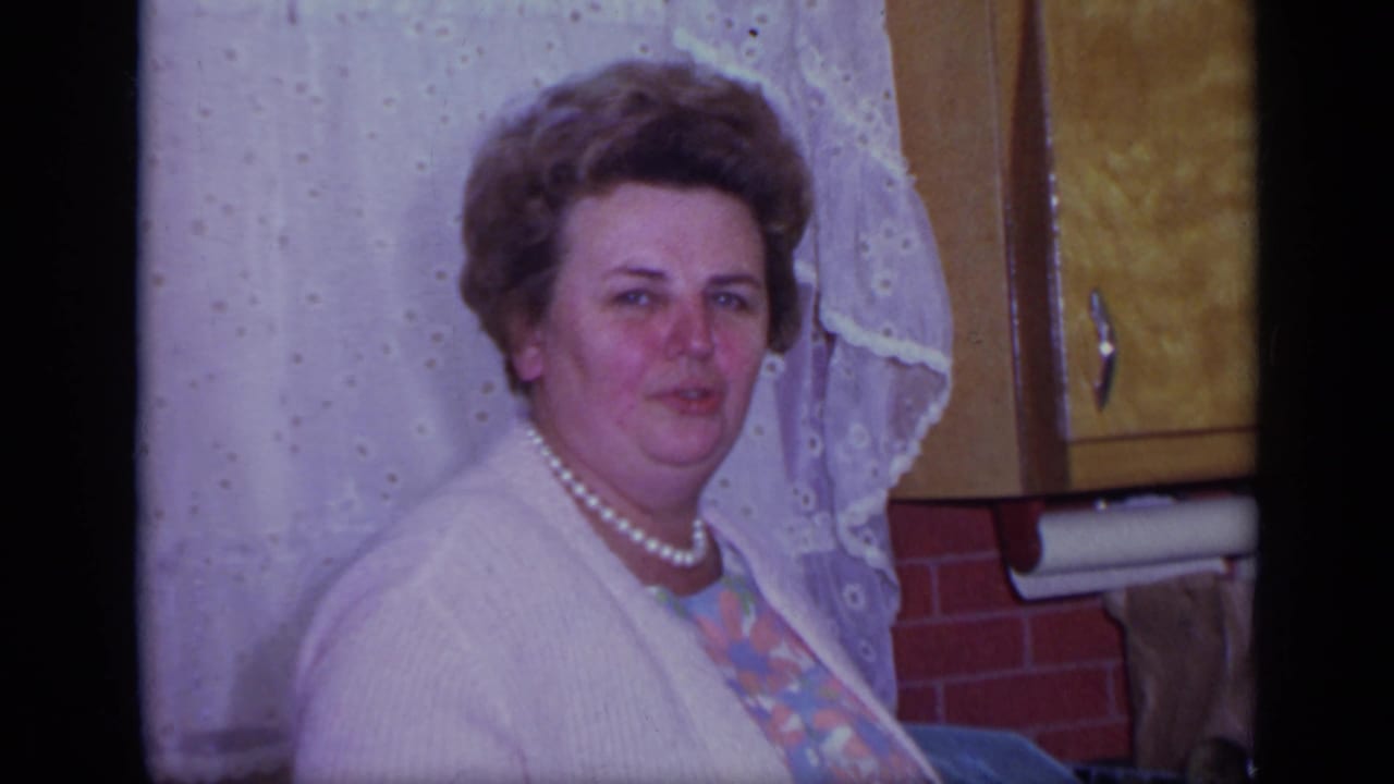 Woman in a white cardigan and pearl necklace seated or standing near a kitchen window.