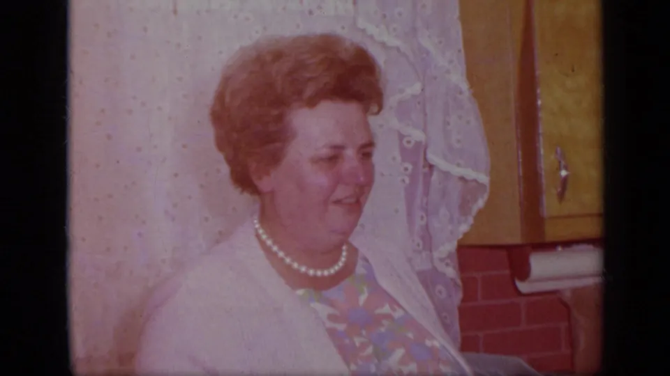The woman turns slightly in profile in the California kitchen, with lace curtains, wood cabinets, and red brick-pattern wall behind her.