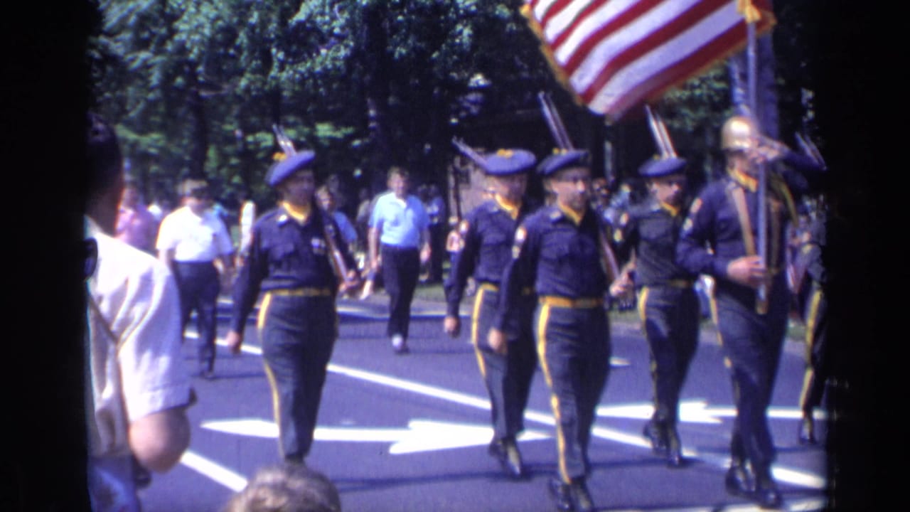 Uniformed marchers in dark blue carry rifles and an American flag along a sunny parade route.