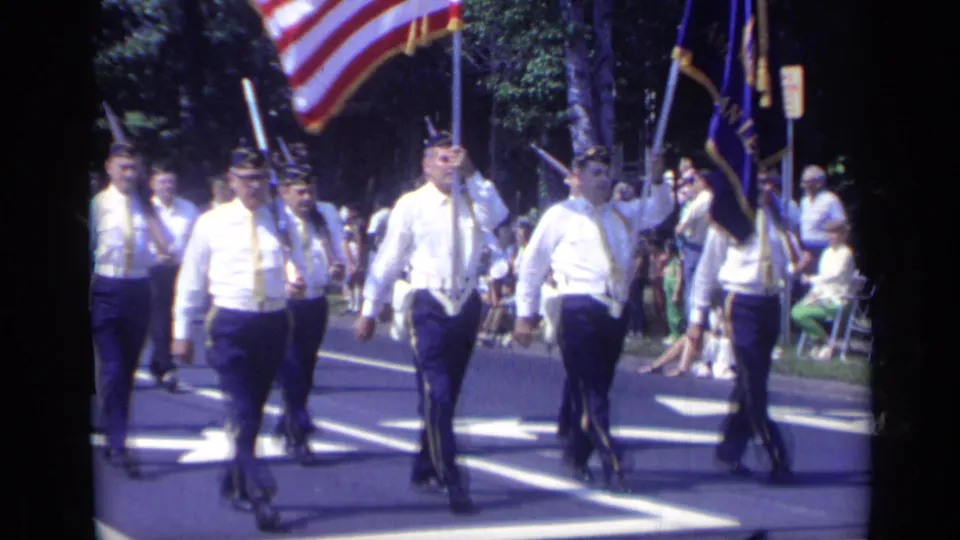 1969 California parade frame showing white-shirted veterans marching with American flags and a blue banner as spectators watch from the sidewalk.