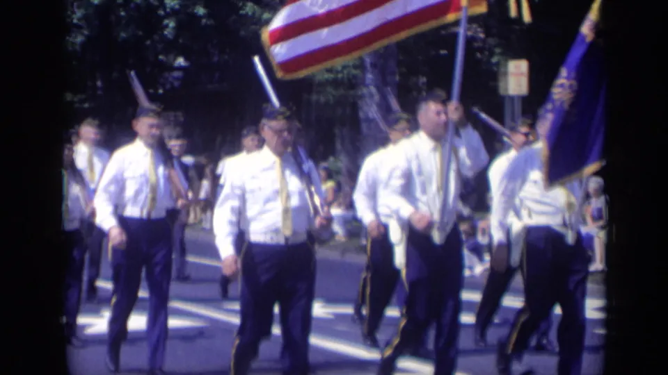 1969 California home-movie frame with veterans in white shirts and dark trousers marching past the camera with flags overhead.