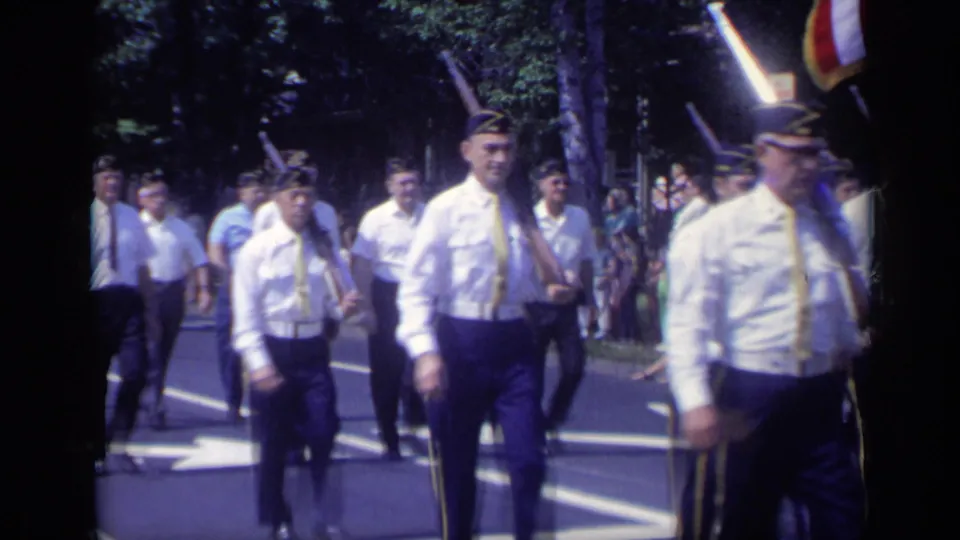 1969 California parade frame showing white-shirted veterans marching along a sunny street with flags, trees, and spectators in view.