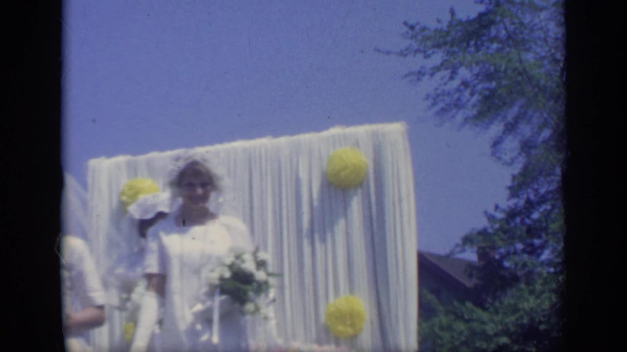 In 1969 California home-movie footage, a young woman in a white dress and veil stands by white drapery and yellow paper decorations on a parade float.