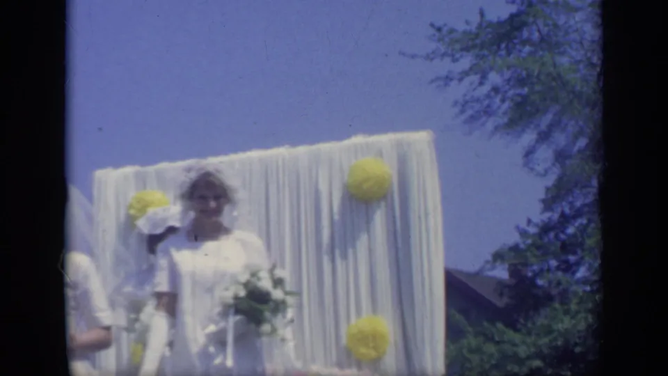 In 1969 California home-movie footage, a young woman in a white dress and veil stands by white drapery and yellow paper decorations on a parade float.