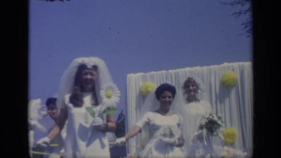 Three young women in white bridal-style outfits and veils ride on a decorated parade float in the California 1969 home-movie clip.