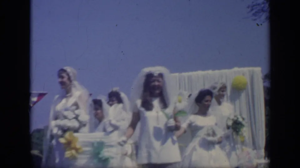 Several bridal-dressed young women in veils and white clothing stand together on a decorated parade float, filmed in 1969 California.