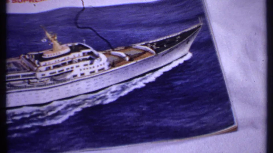 1969 Bahamas home-movie frame showing a printed cruise ship photograph on a light surface, with blue water, wake, and a dark crease near the top.