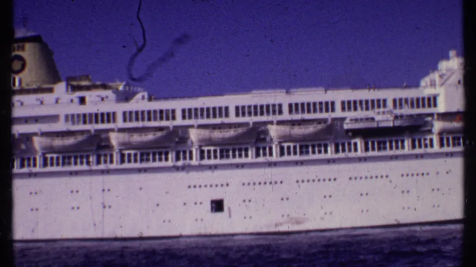 Close home-movie view of the ship's side in the Bahamas, showing lifeboats, a white hull, deck windows, and purple-tinted film grain.