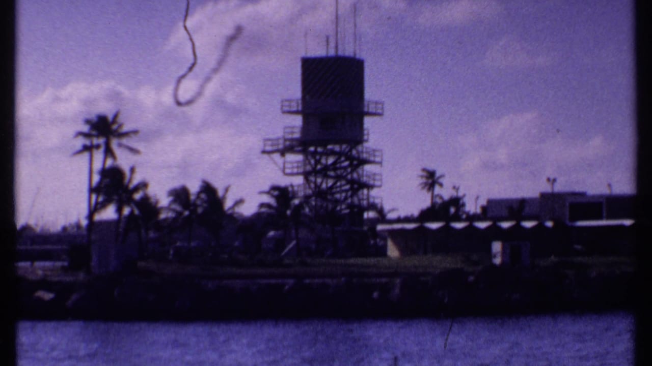 1969 Bahamas home-movie frame of a waterfront tower, palms, low shore buildings, and calm water with 8mm gate edges visible.
