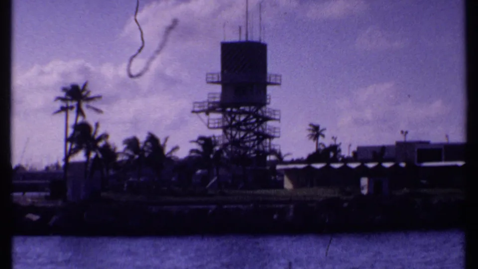 1969 Bahamas home-movie frame of a waterfront tower, palms, low shore buildings, and calm water with 8mm gate edges visible.