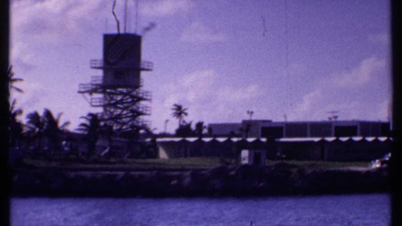 1969 Bahamas frame showing a tower on a tropical waterfront, with palms, low buildings, and blue-purple 8mm color cast.