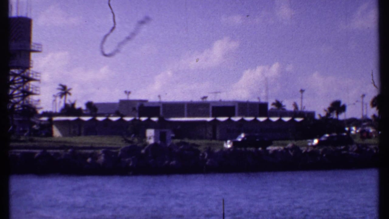 1969 Bahamas shoreline frame with low modern building, parked cars, palms, water, and the edge of a tower entering from the left.