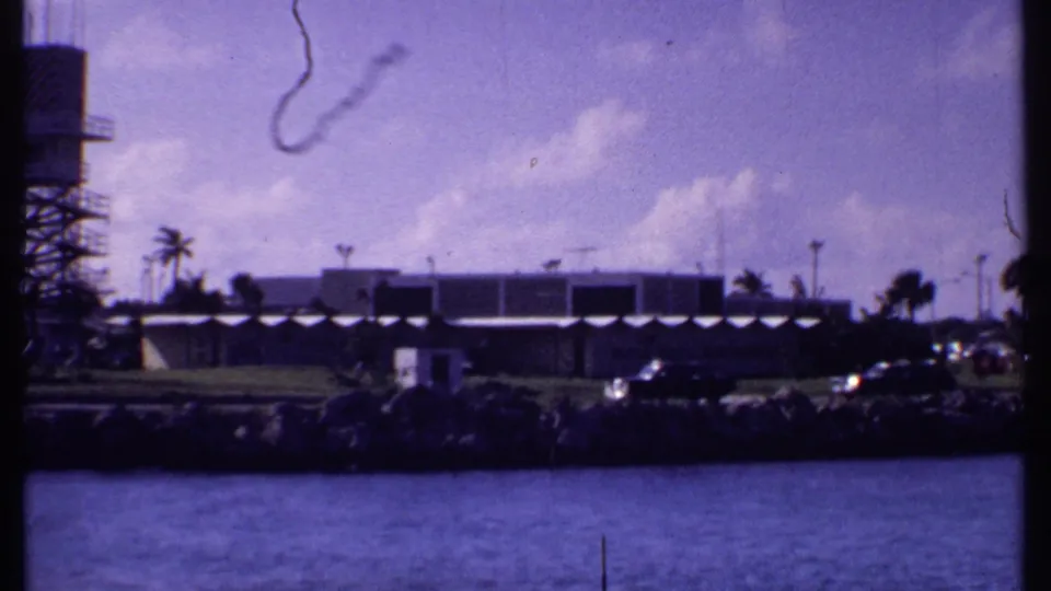 1969 Bahamas shoreline frame with low modern building, parked cars, palms, water, and the edge of a tower entering from the left.