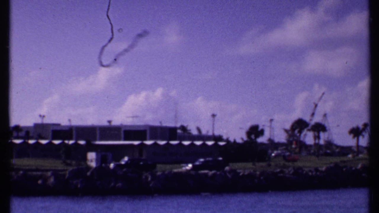 1969 Bahamas home-movie frame of waterfront buildings and palms beneath bright clouds, with a thin contrail-like streak in the sky.