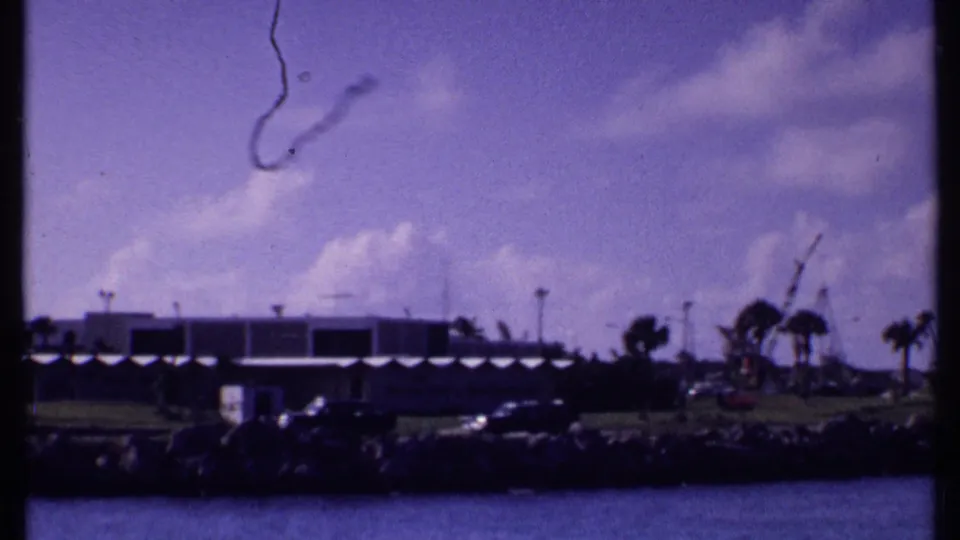 1969 Bahamas home-movie frame of waterfront buildings and palms beneath bright clouds, with a thin contrail-like streak in the sky.