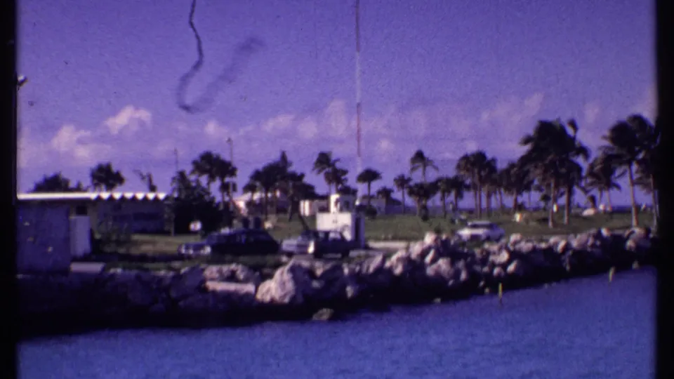 1969 Bahamas waterfront view with a rocky jetty, palms, low buildings, parked cars, and blue water in the foreground.