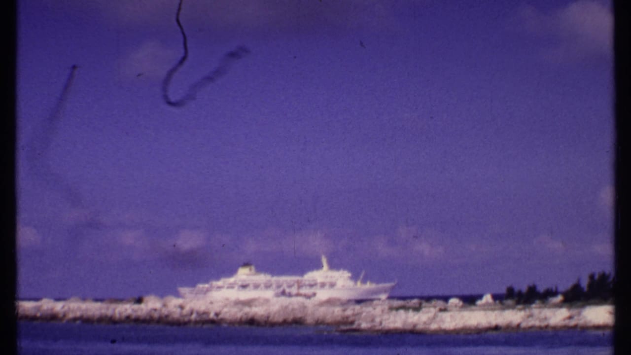 A distant white cruise ship sits behind the breakwater in this Bahamas home-movie frame, with shoreline rocks and open water visible.