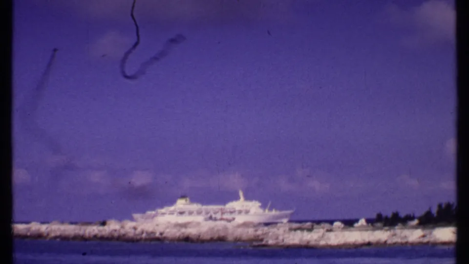 A distant white cruise ship sits behind the breakwater in this Bahamas home-movie frame, with shoreline rocks and open water visible.
