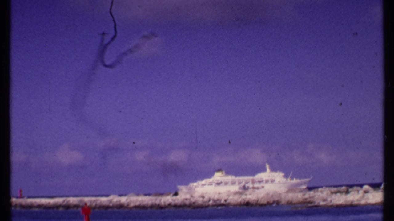 1969 Bahamas 8mm frame showing a white cruise ship behind a breakwater with a dark smoke-like trail in the sky.