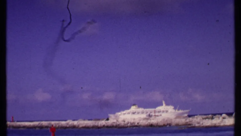 1969 Bahamas 8mm frame showing a white cruise ship behind a breakwater with a dark smoke-like trail in the sky.