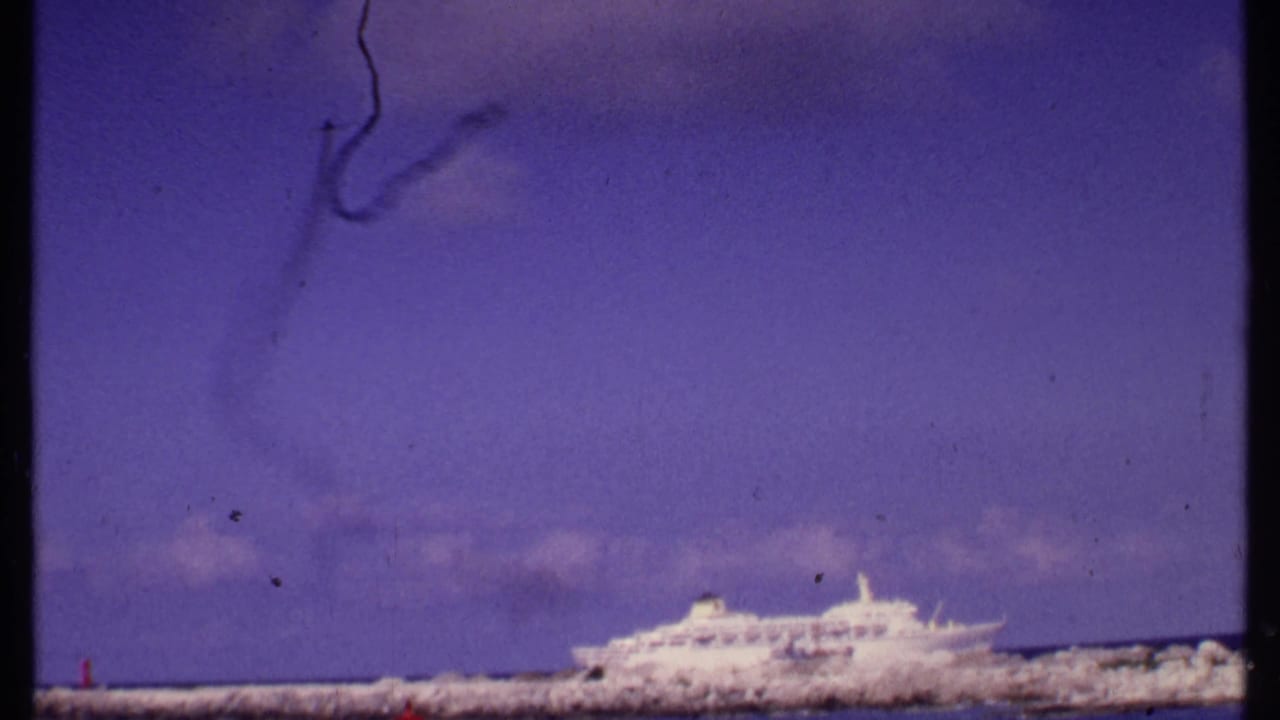1969 Bahamas harbor view with a white cruise ship, low breakwater, blue-purple sky, and visible 8mm film wear.