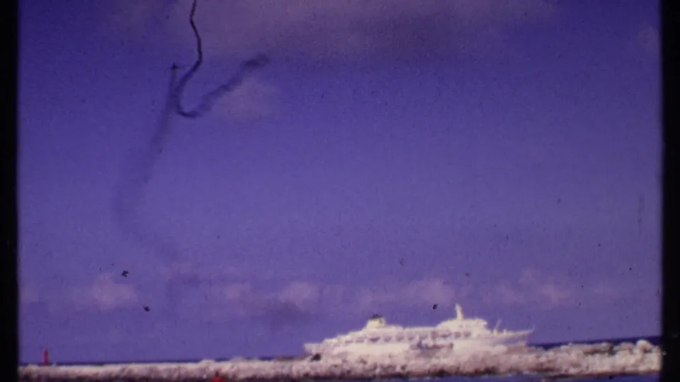 1969 Bahamas harbor view with a white cruise ship, low breakwater, blue-purple sky, and visible 8mm film wear.